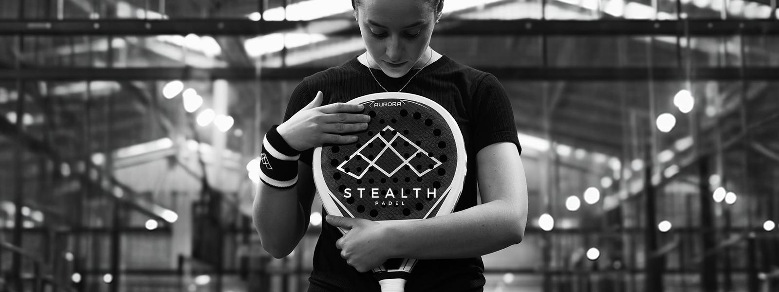 Eine Padel-Spielerin hält den Stealth Aurora Padelschläger vor der Brust in einer Padel-Halle; hochwertiges Schwarz-Weiß-Lifestyle-Foto der Stealth-Kollektion.